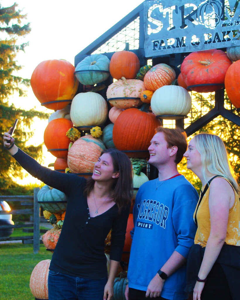 Young adults take a group selfie​ in front of a Strom's Farm & Bakery sign.
