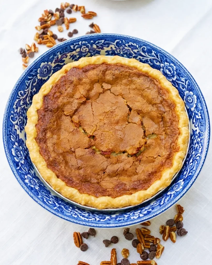 A whole homemade chocolate pecan pie featuring our made-from-scratch pastry crust. The pie is presented in a blue and white floral dish and surrounded by scattered pecans and chocolate chips on a white tablecloth at Strom's Farm & Bakery in Guelph, Ontario.