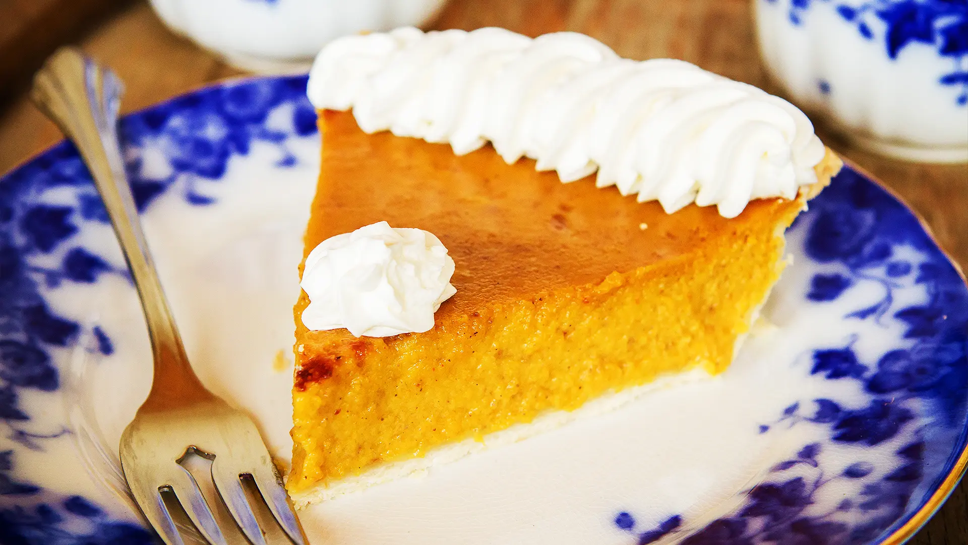 A slice of homemade pumpkin pie featuring our made-from-scratch pastry and farm-grown pumpkin puree. The slice is topped with whipped cream and served on a blue and white floral plate with a silver fork at Strom's Farm and Bakery in Guelph, Ontario.